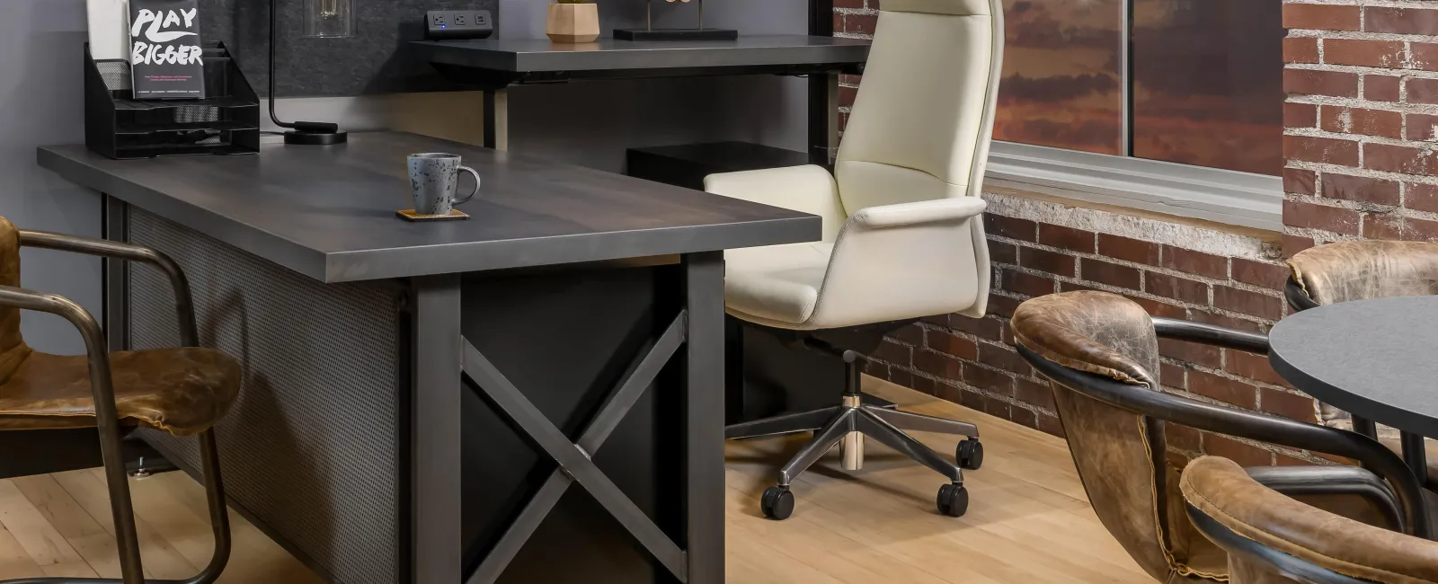 Modern office space with a large dark metal desk, white leather chair, and brown leather seating against brick walls.