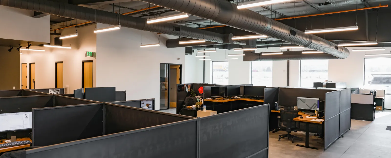 Modern open office space with cubicles, computer workstations, exposed ductwork, and bright overhead lighting.