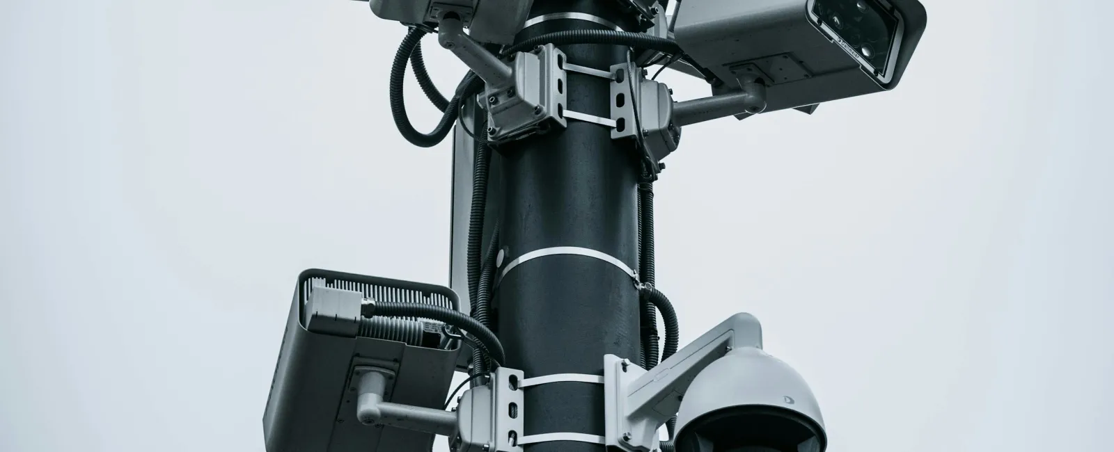 Multiple security cameras mounted on a tall black pole under a cloudy sky for surveillance.