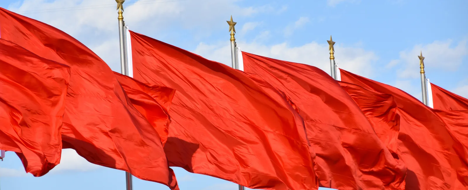 Row of bright red flags waving on poles against a blue sky with scattered clouds.