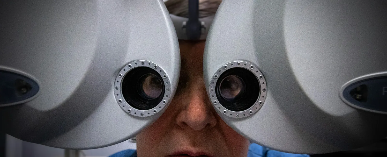 Person undergoing eye exam looking through a phoropter optical eye testing device in a clinic.