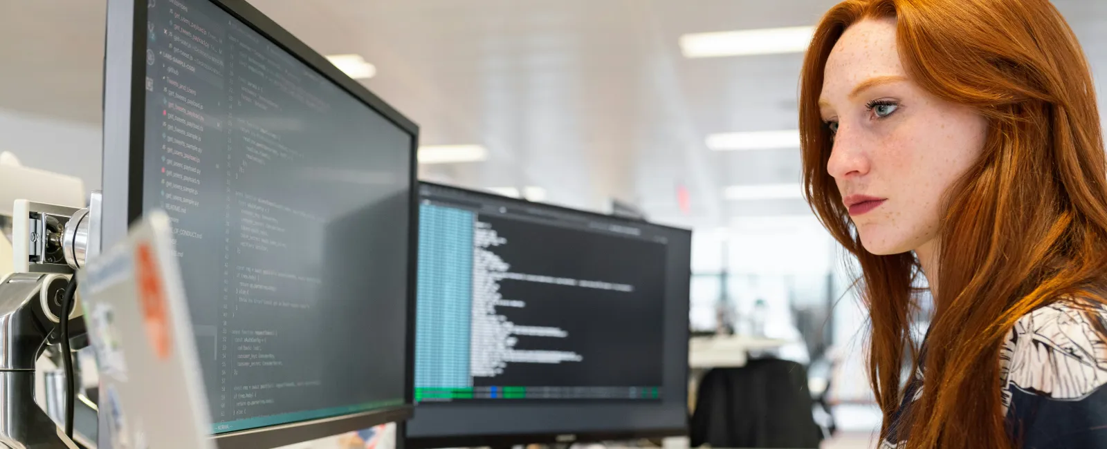Red-haired woman focused on coding using dual monitors and a laptop at a bright office desk.