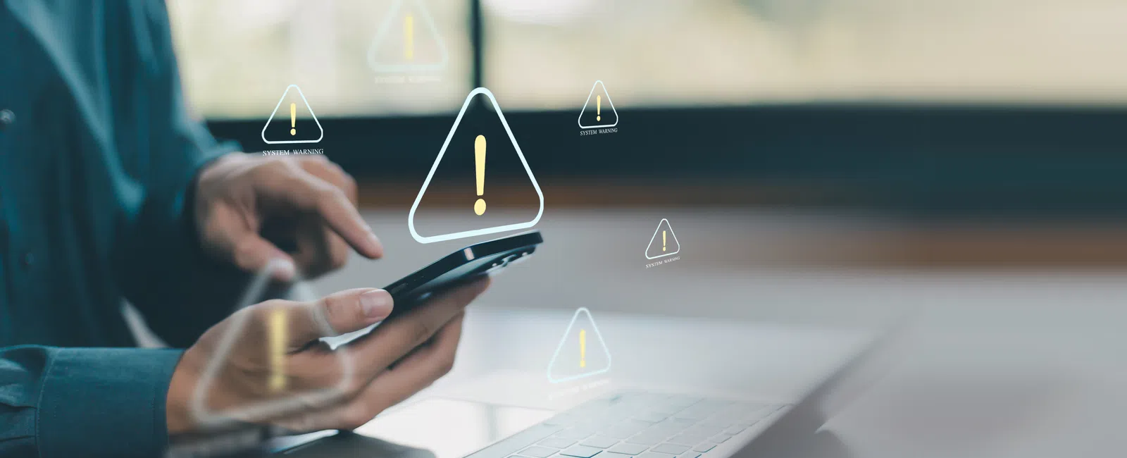 Person using smartphone with digital warning icons floating over laptop keyboard in office setting.