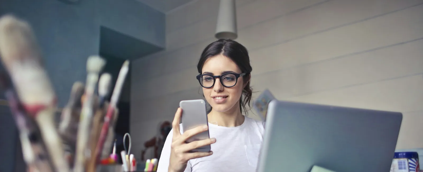 Young woman with glasses using smartphone and laptop in a creative workspace with art supplies visible.