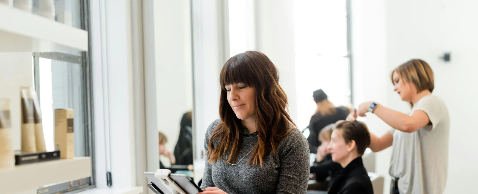 Woman using a tablet near a salon window while hairstylists work on clients in the background