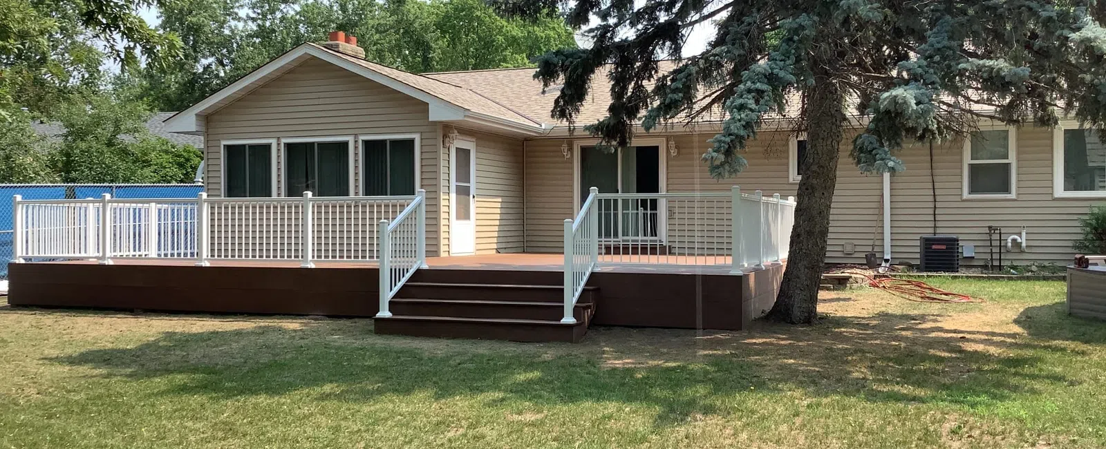 Spacious backyard with deck and railing attached to a single-story beige house with large windows and trees.