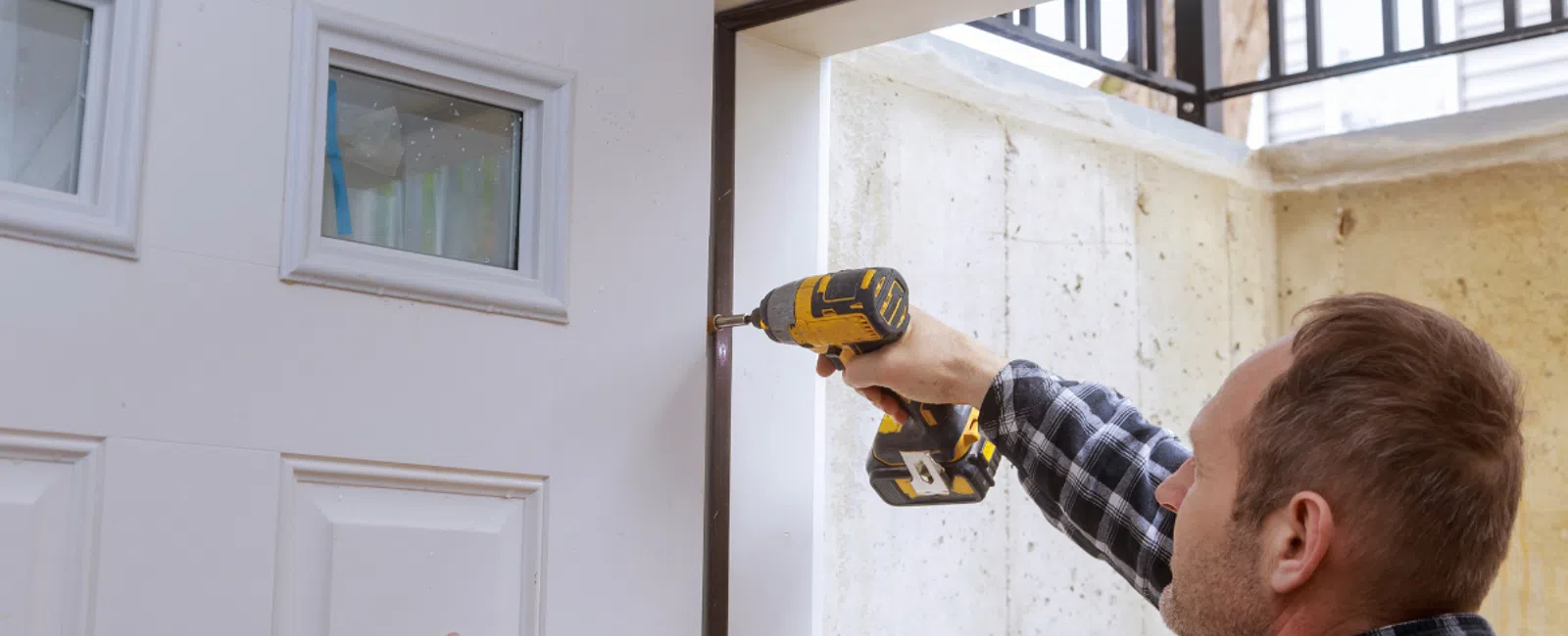 Man installing a white door using a yellow cordless drill in a stone-walled entryway.