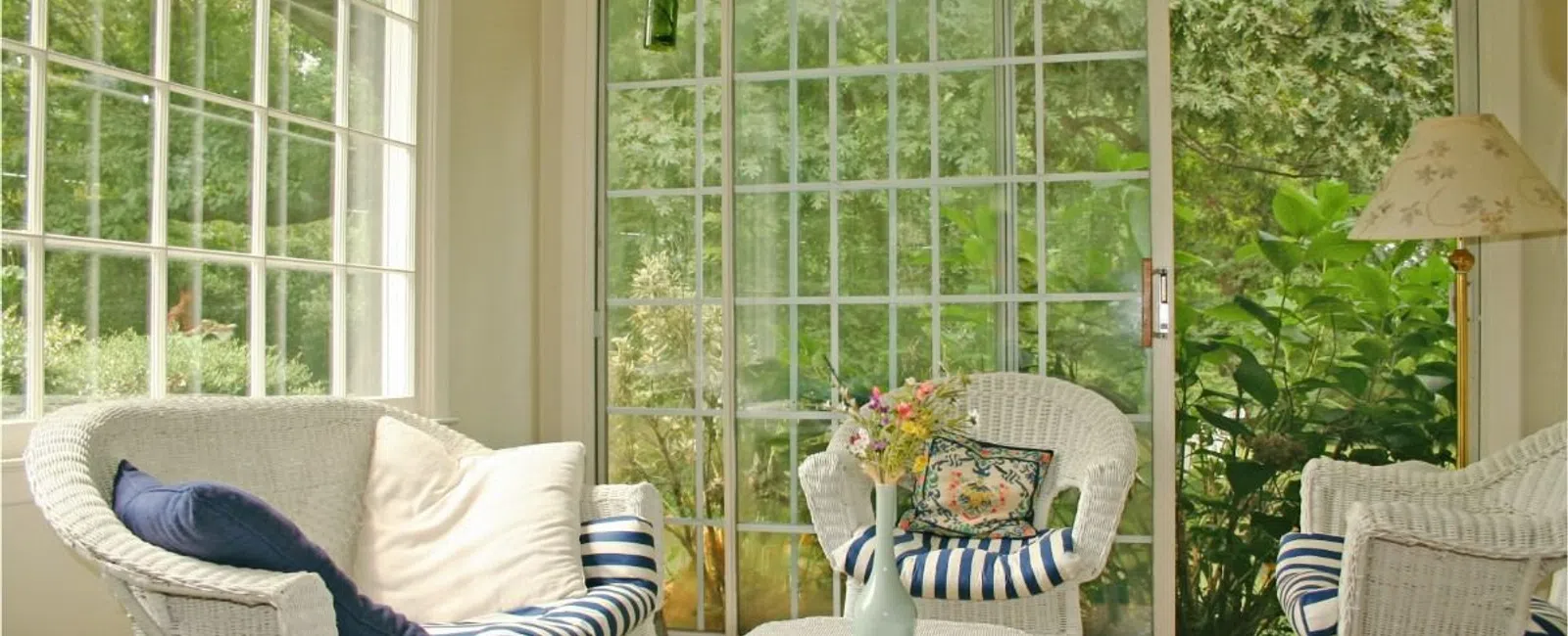 Bright sunroom with white wicker furniture, striped cushions, floral pillows, and large sliding glass doors showing greenery.