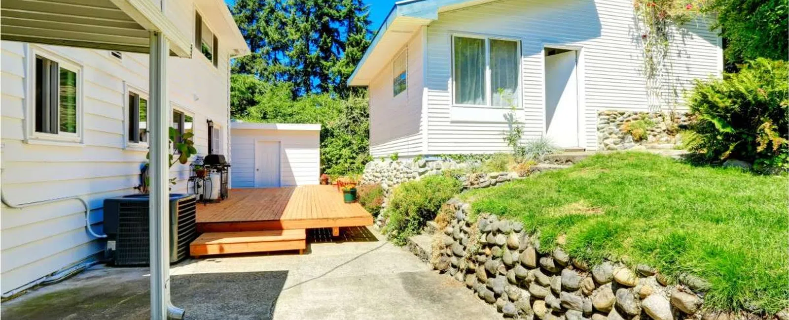 Sunny backyard with white houses, wooden deck, stone retaining wall, and green lawn under clear blue sky.