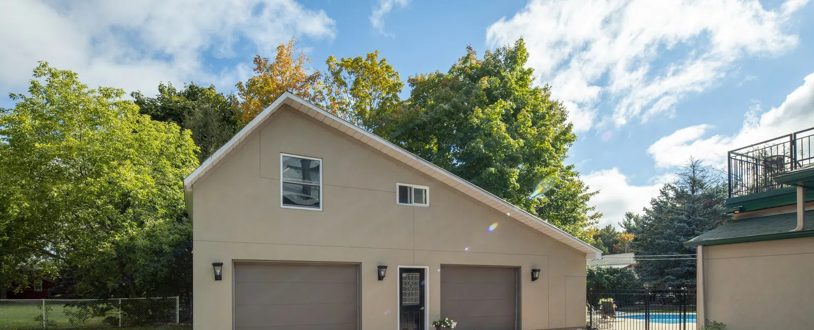 Modern beige three-car garage with paved driveway surrounded by green trees under a blue cloudy sky