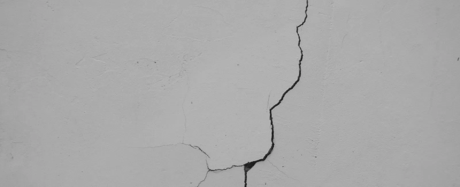 Close-up of a gray concrete wall with prominent vertical and horizontal cracks indicating damage.