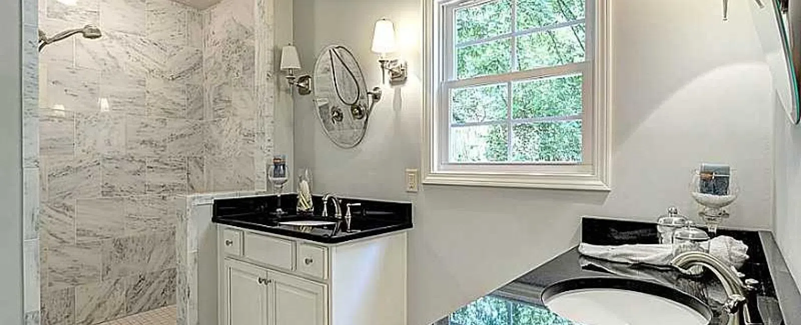 Modern bathroom with dual black granite sinks, white cabinetry, marble walk-in shower, and window with natural light.