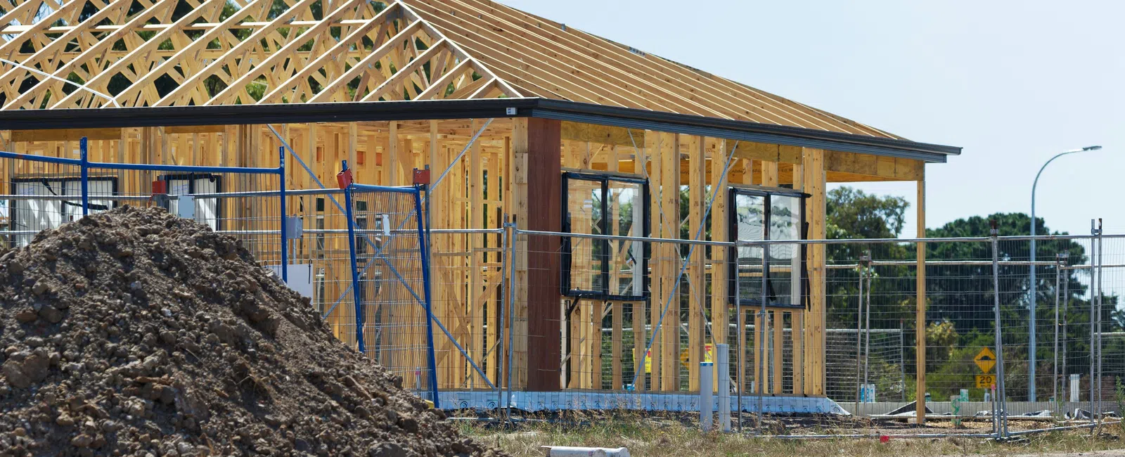 Wooden frame structure of a house under construction on a sunny day with a dirt pile in the foreground.