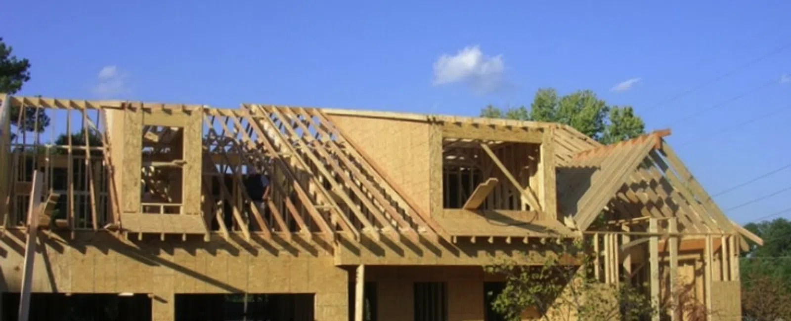 Partially framed house under construction with wooden beams and plywood sheathing against a blue sky.