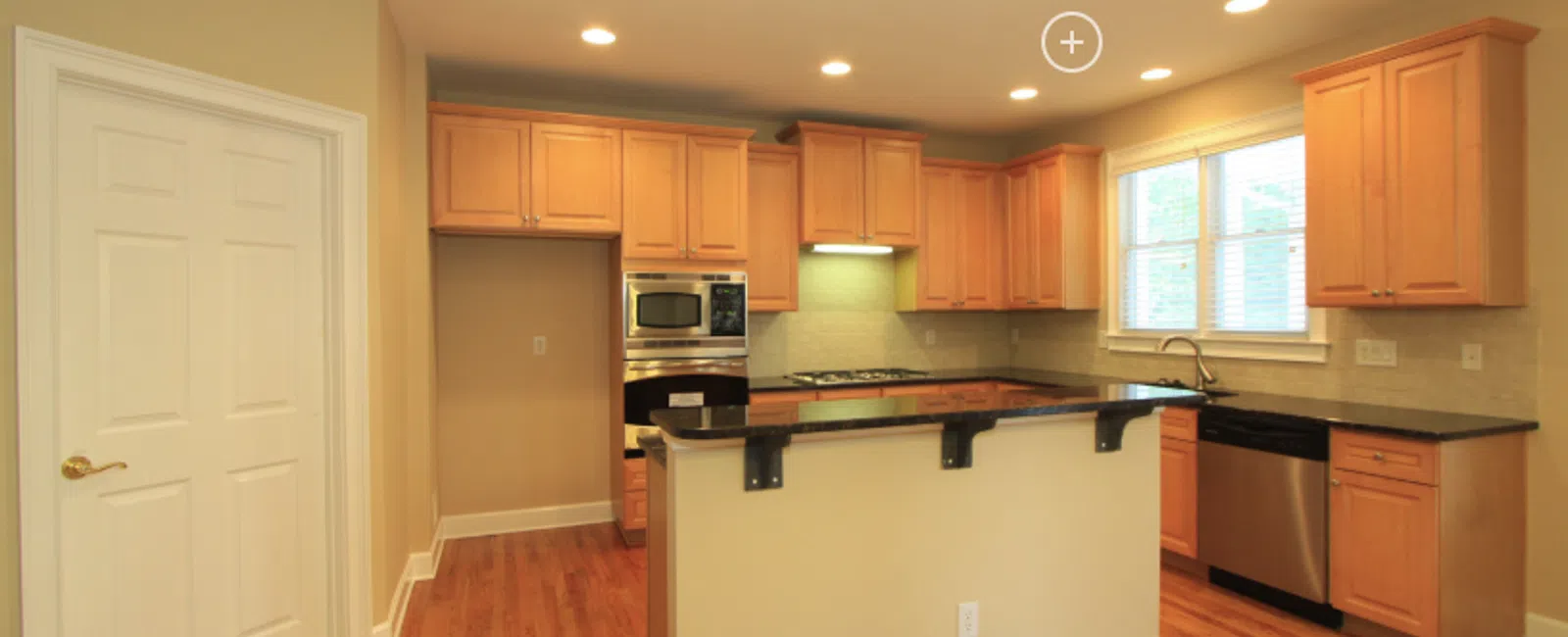 Bright kitchen with wooden cabinetry, black countertops, stainless steel appliances, and hardwood floors in a modern home.