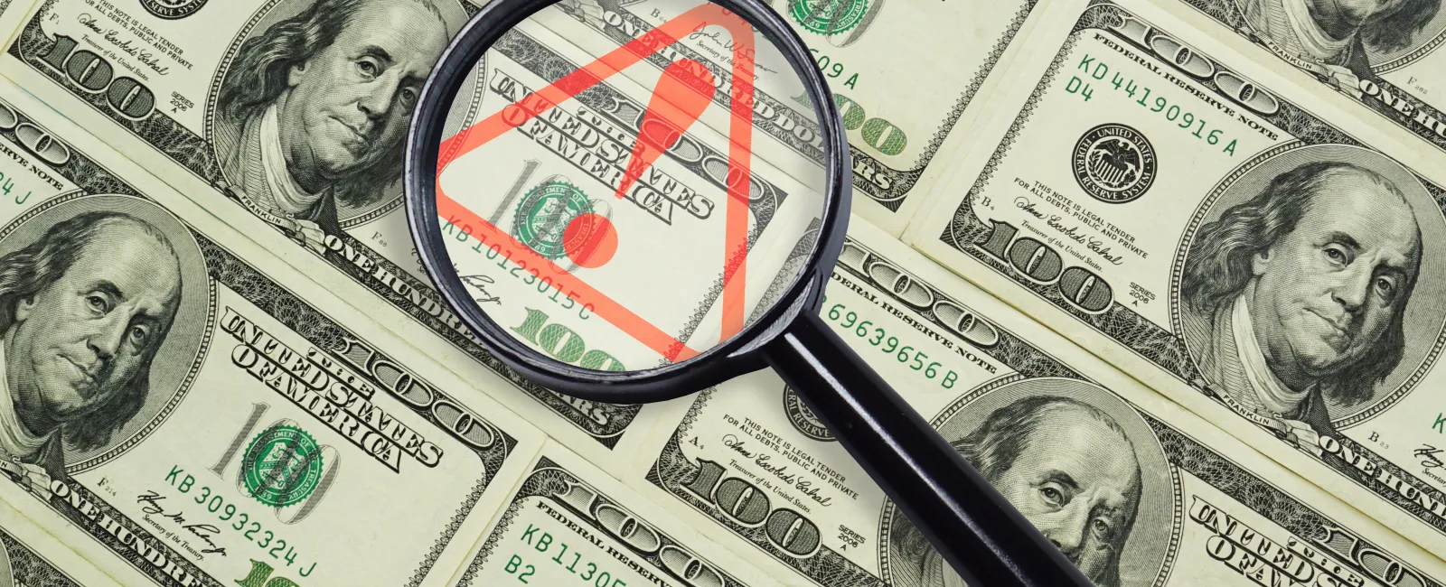 Magnifying glass highlighting a red warning symbol over scattered US hundred-dollar bills depicting financial risk.