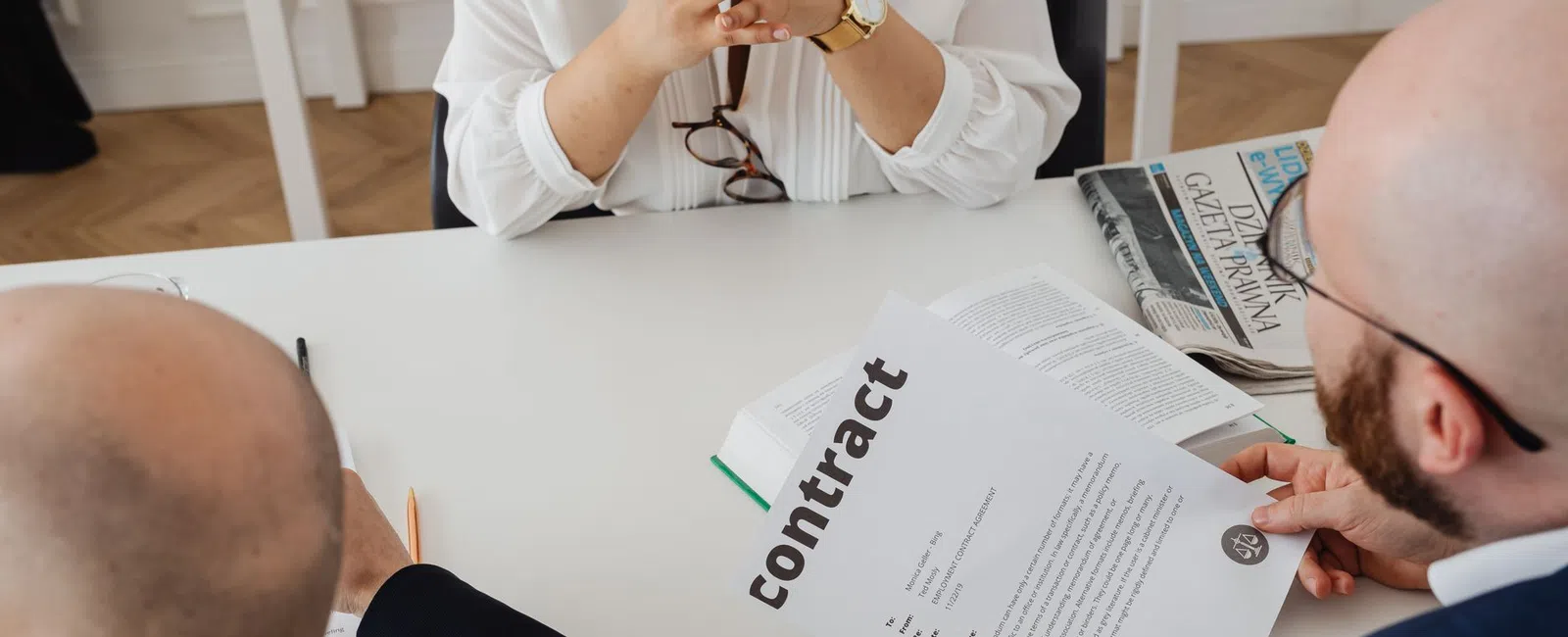 Two men reviewing a contract document across a desk from a woman in a white blouse with folded hands.