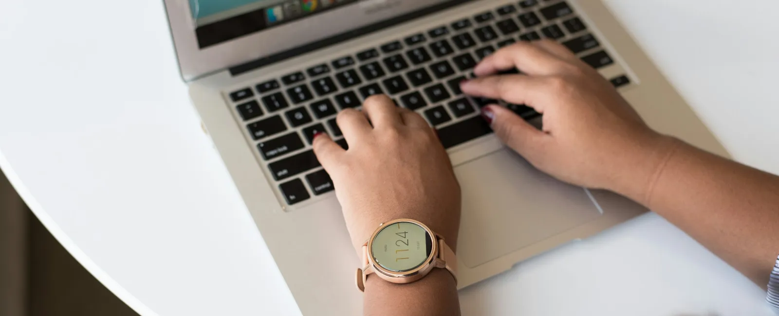 Person typing on a laptop keyboard wearing a rose gold smartwatch on a white round table.
