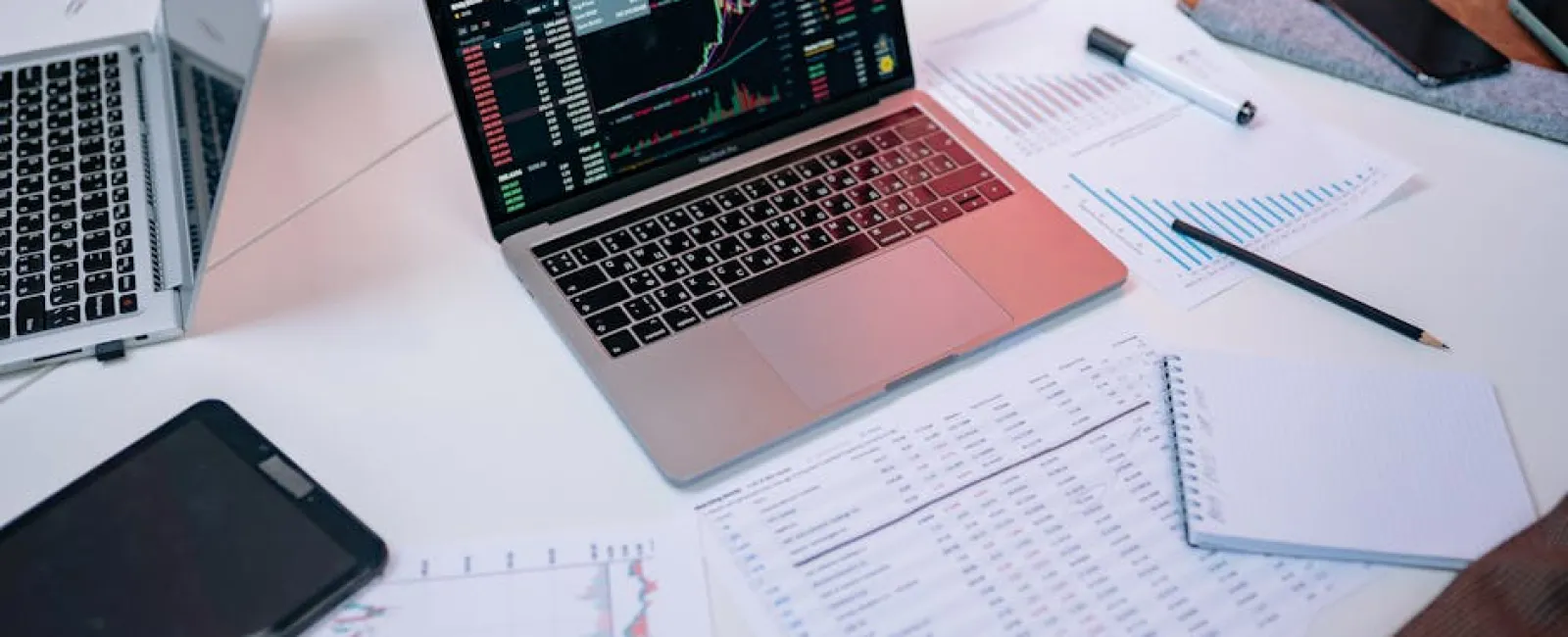 Laptop displaying stock market charts surrounded by financial documents, notebooks, and a smartphone on a white table