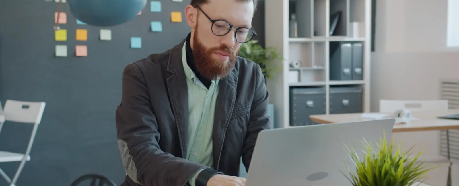 Man with glasses working on laptop at desk in modern office with sticky notes on wall and green plant