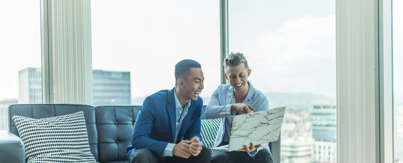 Two business professionals smiling and discussing on a sofa with a laptop in a modern office lounge.