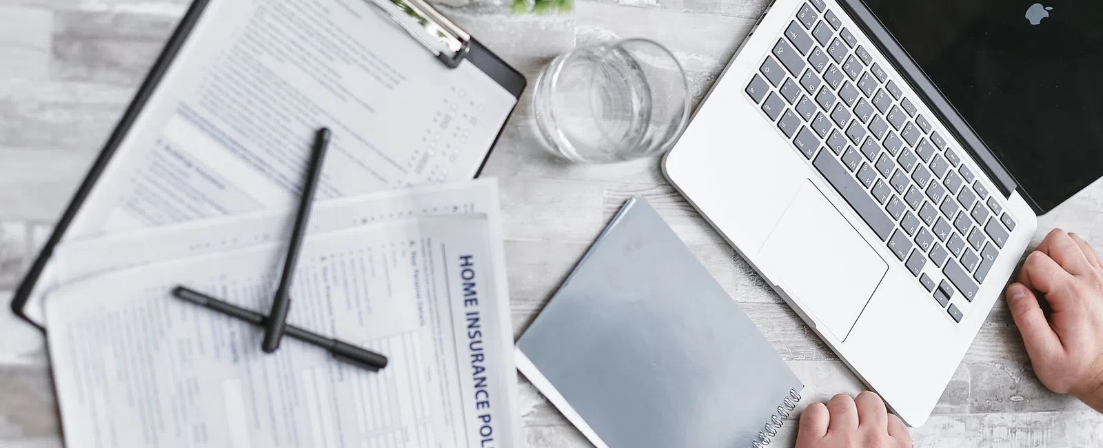 Person working at a desk with a laptop, home insurance policy documents, notebook, and glass of water.