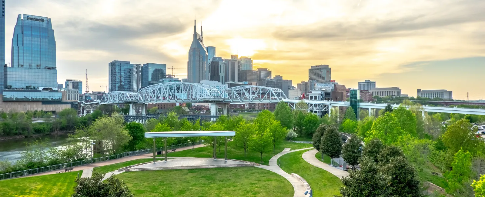 Sunset over Nashville skyline with green park, river, and pedestrian bridge in foreground under cloudy sky