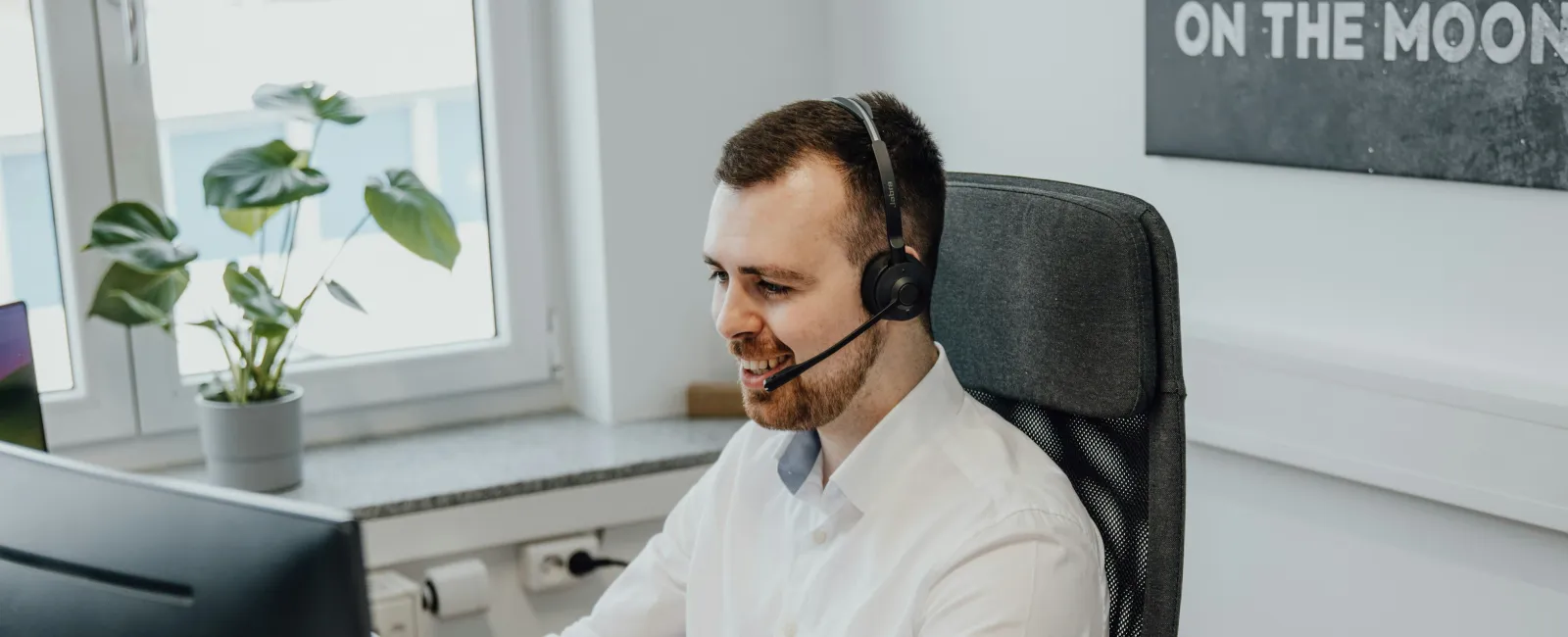 Man in white shirt wearing headset working on a computer in a bright modern office with a plant nearby.