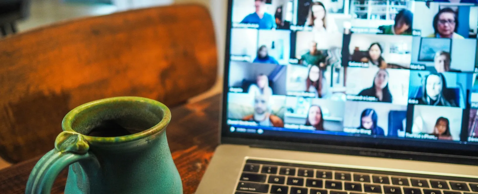 Laptop showing a virtual meeting with multiple participants next to a green ceramic coffee mug on a wooden table.