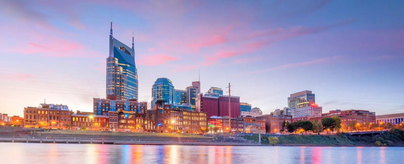Nashville city skyline at sunset with colorful reflections on the river and glowing city lights.