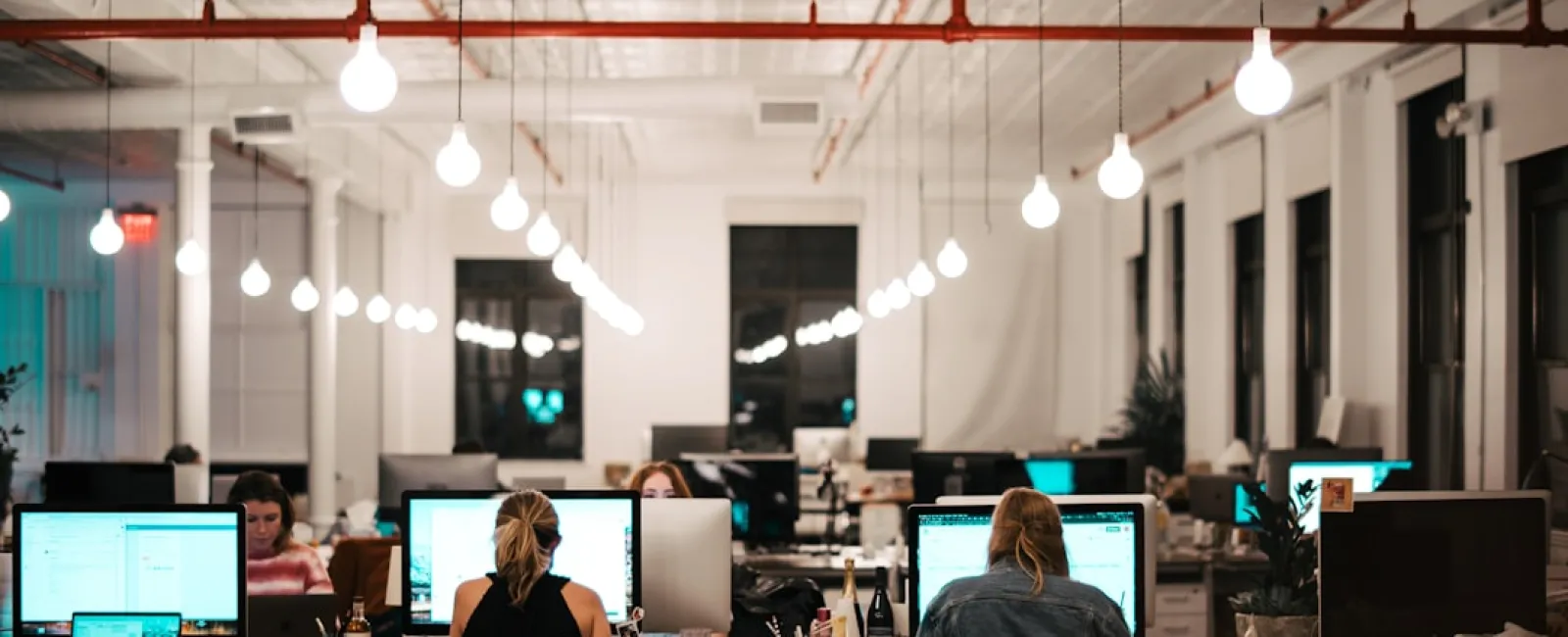 Modern open-plan office with workers at desks using computers under hanging light bulbs at night.