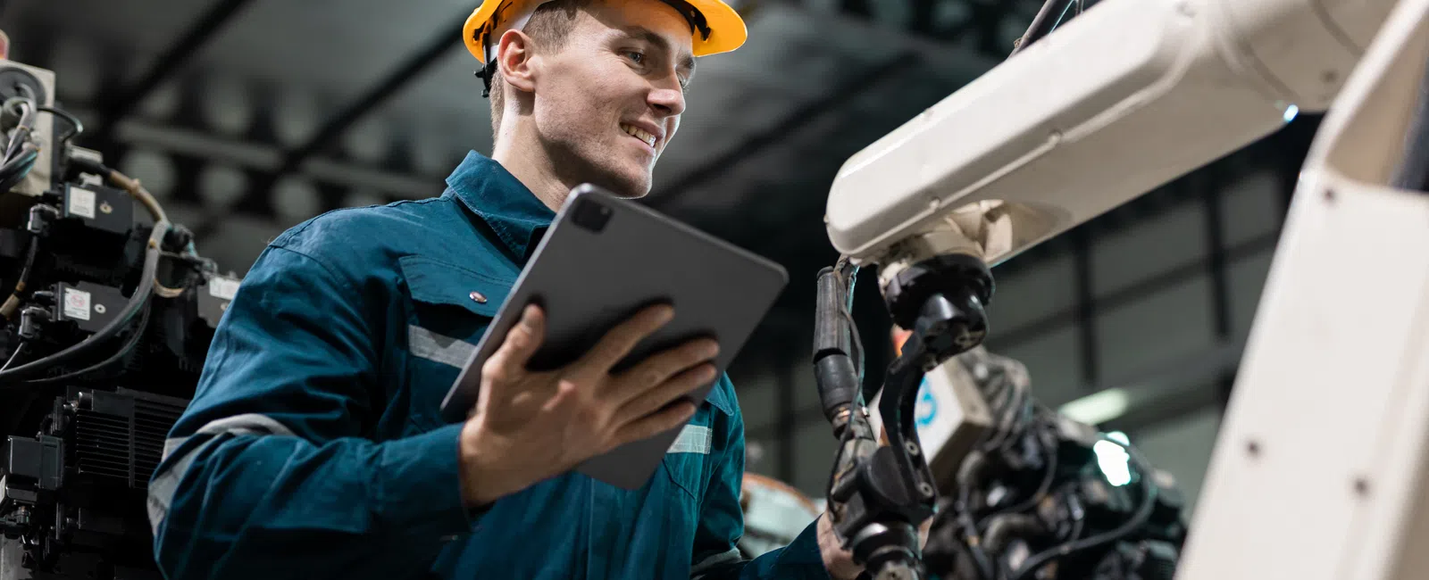 Engineer in protective gear using tablet to operate robotic arm in an industrial factory setting.