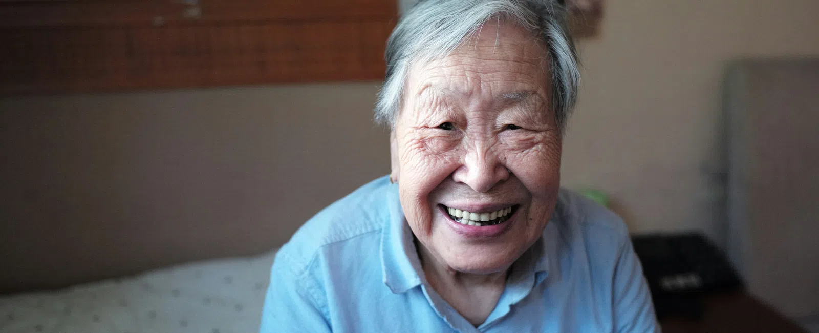 Elderly Asian woman smiling warmly while sitting on a bed in a softly lit room, wearing a blue shirt.