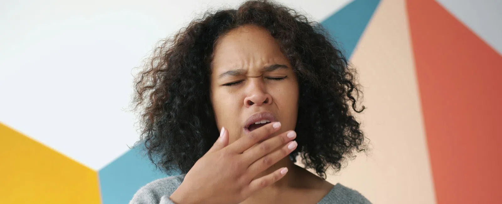 Young woman yawning with eyes closed against a colorful geometric wall background.