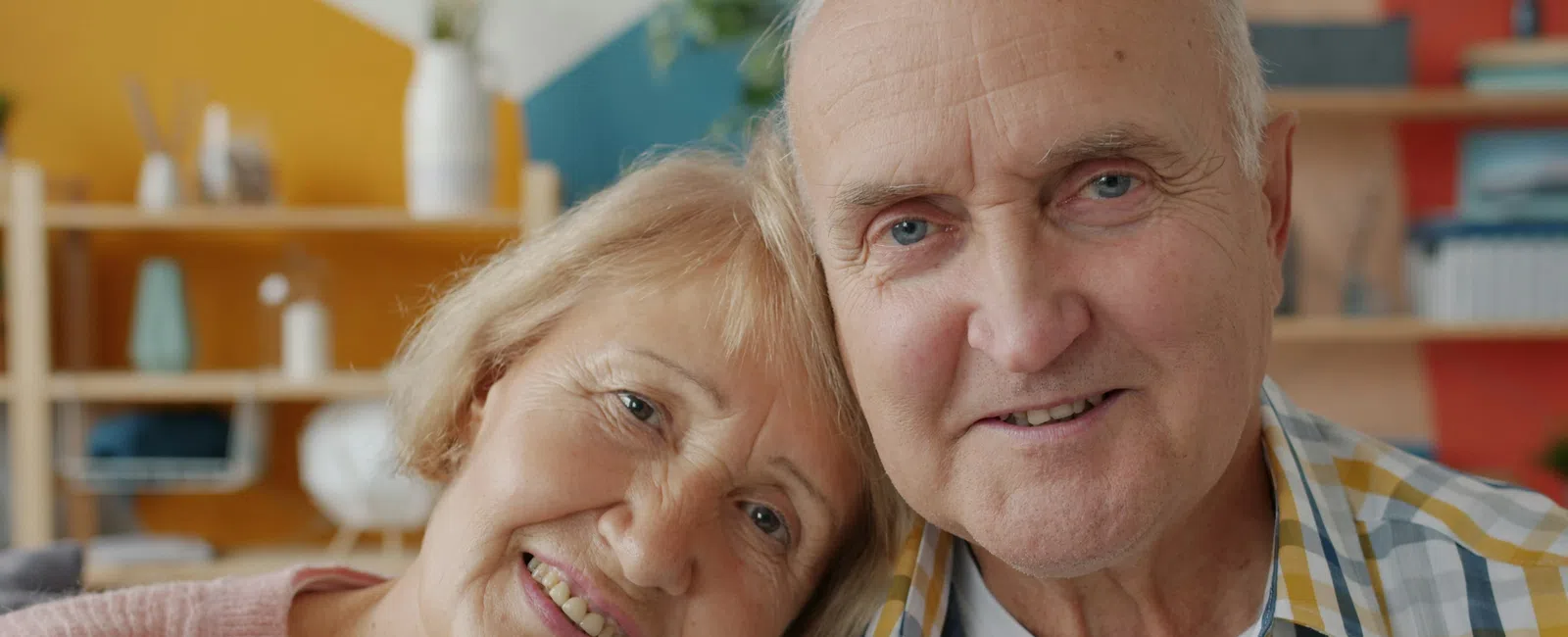 Smiling elderly couple sitting closely indoors with warm lighting and colorful background elements.