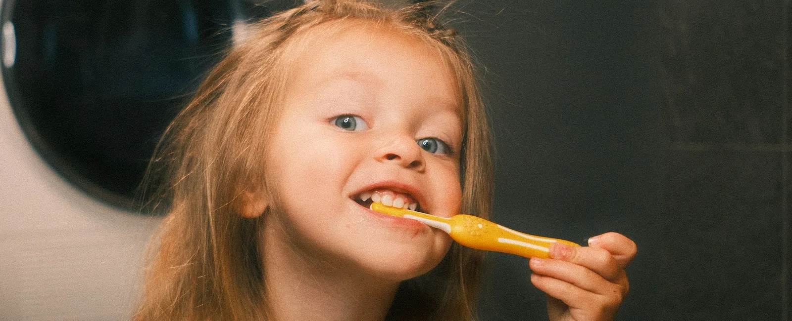 Young child brushing teeth with yellow toothbrush in bathroom near washing machine and mirror
