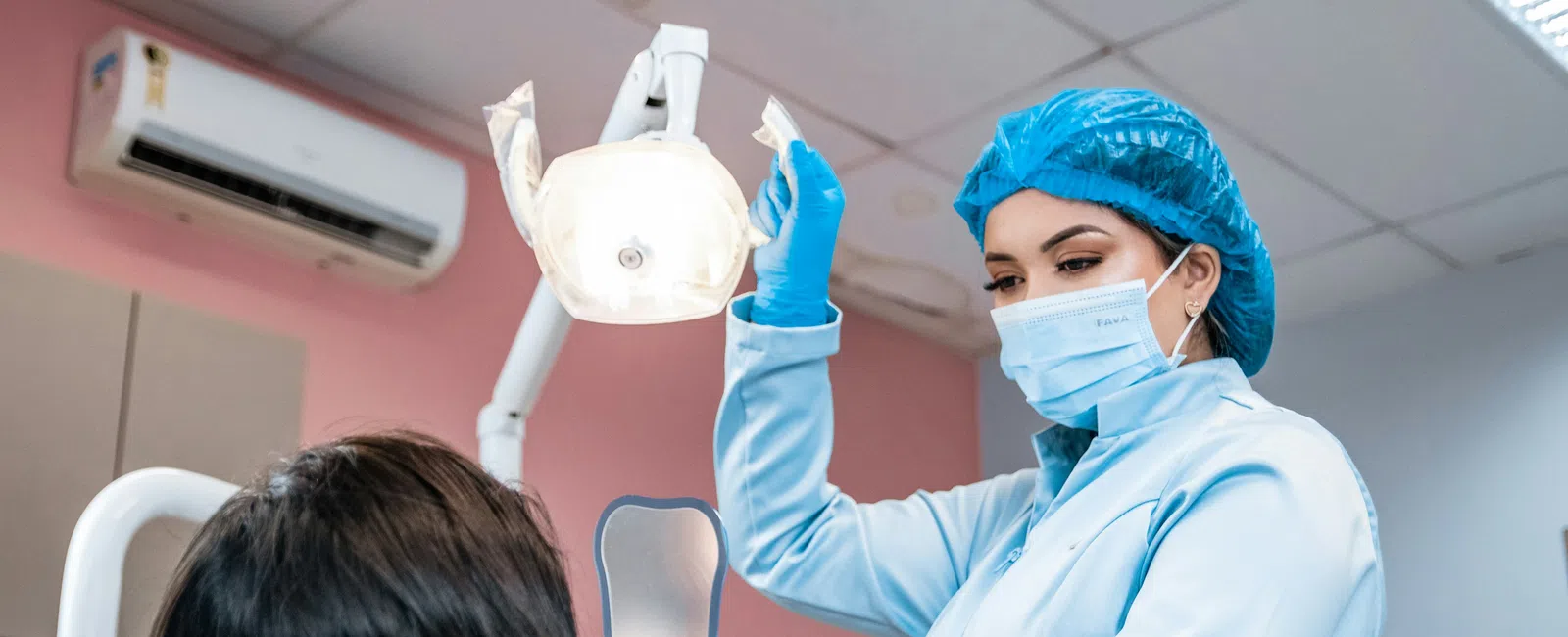 Dentist in protective gear preparing dental light while patient holds a mirror in the dental clinic chair.