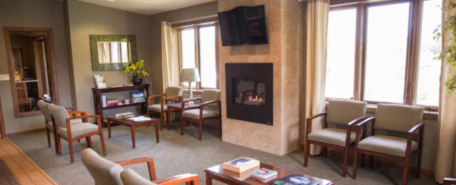 Cozy waiting room with cushioned chairs, a fireplace, flat-screen TV, and natural light from large windows.