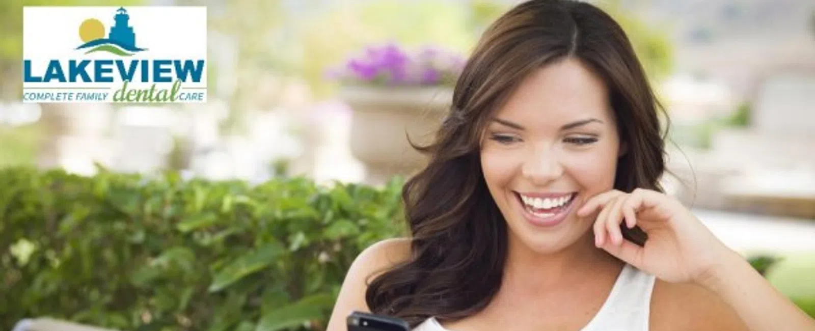 Happy young woman smiling while looking at her smartphone outdoors on a park bench with greenery background