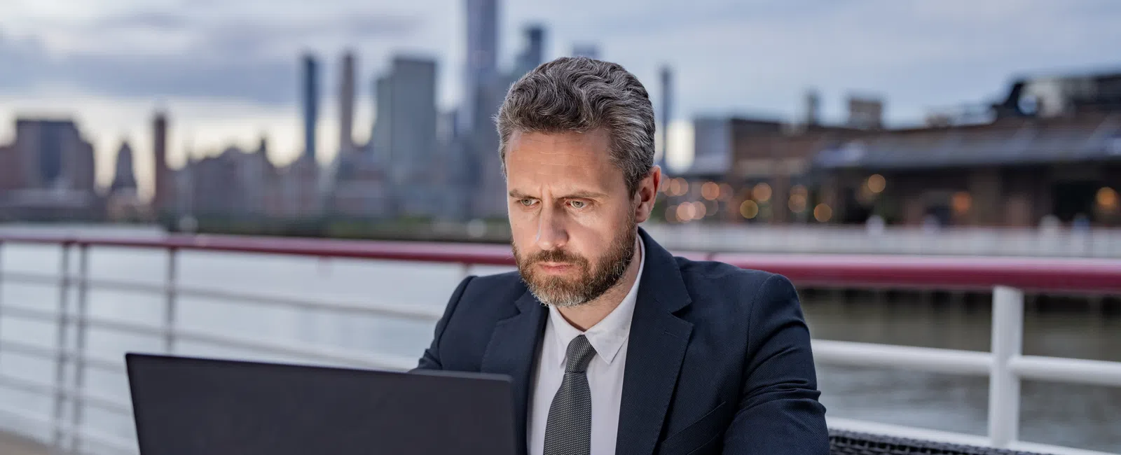 Focused businessman in suit working on laptop at outdoor table with city skyline in background at dusk.