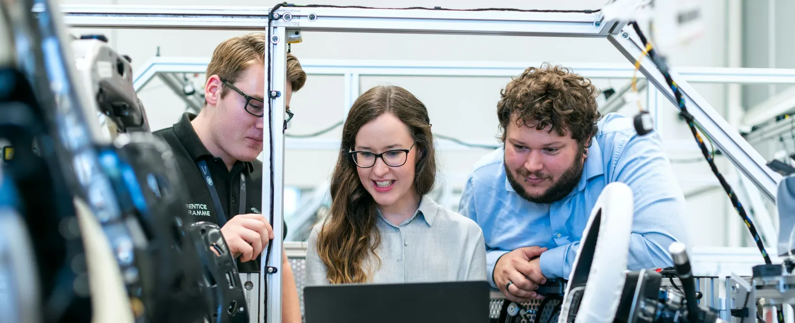 Three engineers collaborate on a laptop inside a car prototype in a modern technical workshop environment