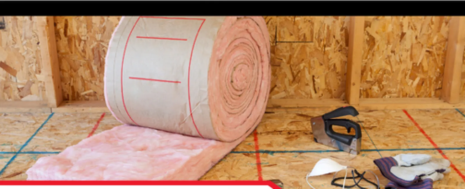 Roll of pink fiberglass insulation with safety gear and staple gun on plywood subfloor in attic framing.