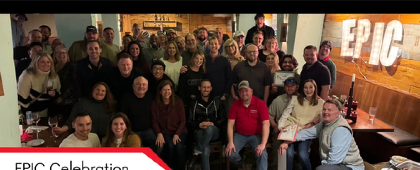 Large group of diverse people smiling and posing for a celebratory photo inside a cozy venue with EPIC sign.