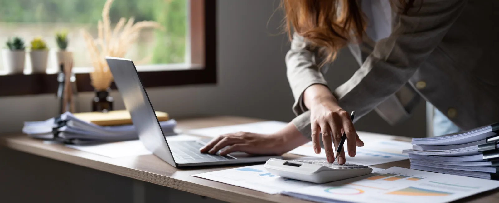 Person using calculator and laptop while working with financial documents on a wooden desk near window.
