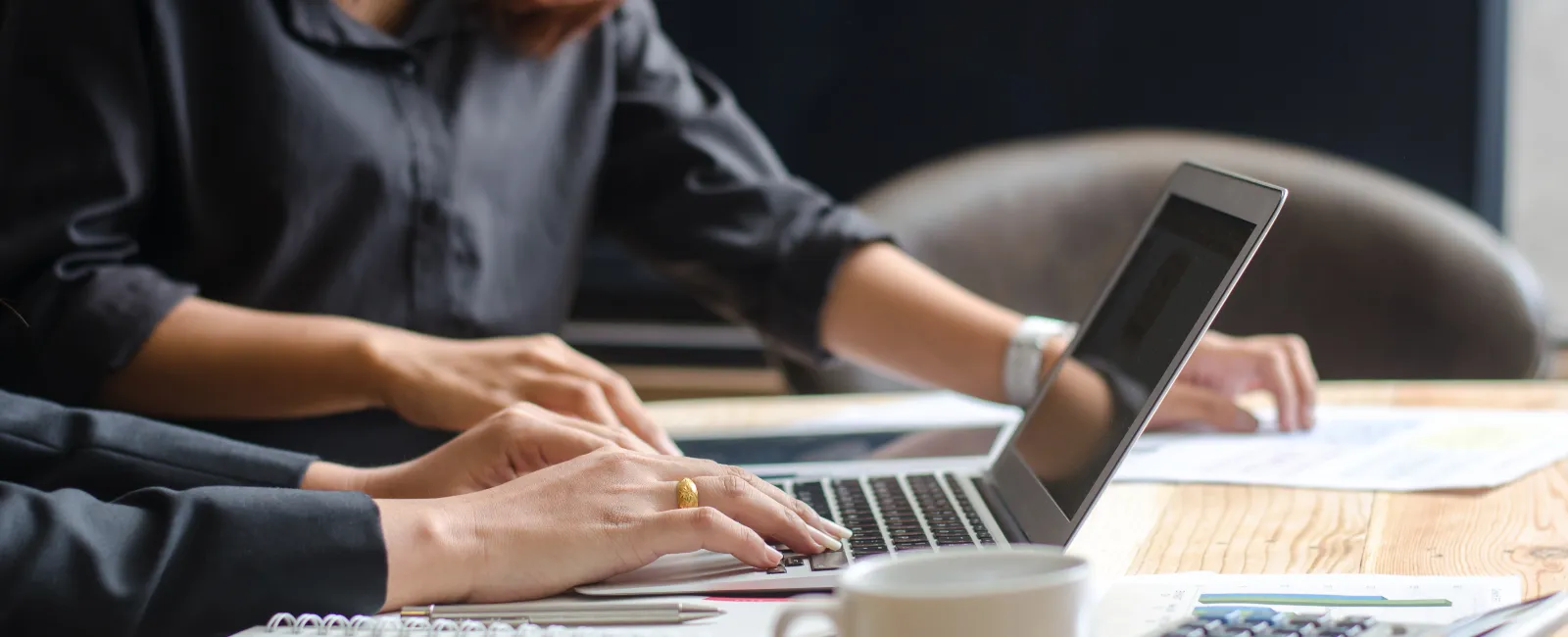 Two professionals working at a wooden desk with laptop, documents, calculator, and coffee cup in a focused office setting.