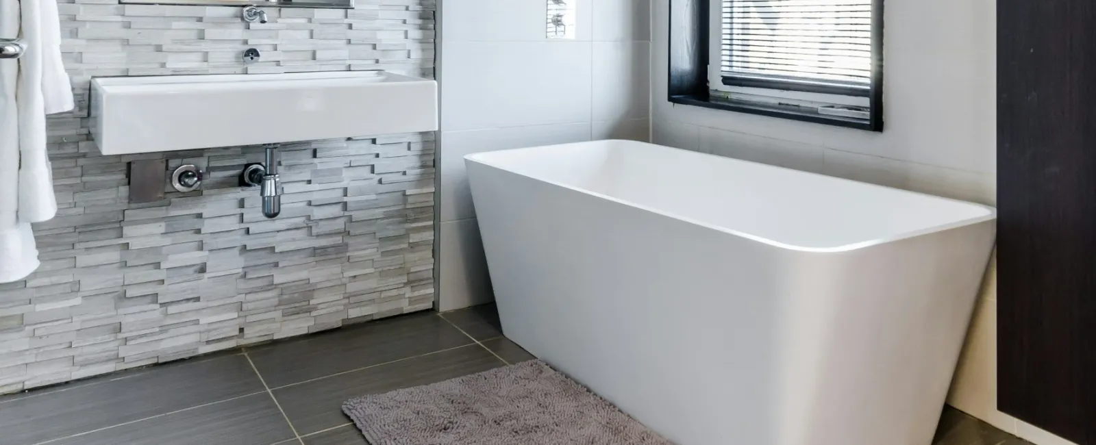 Modern bathroom with freestanding white tub, wall-mounted sink, textured tile wall, and window blinds.