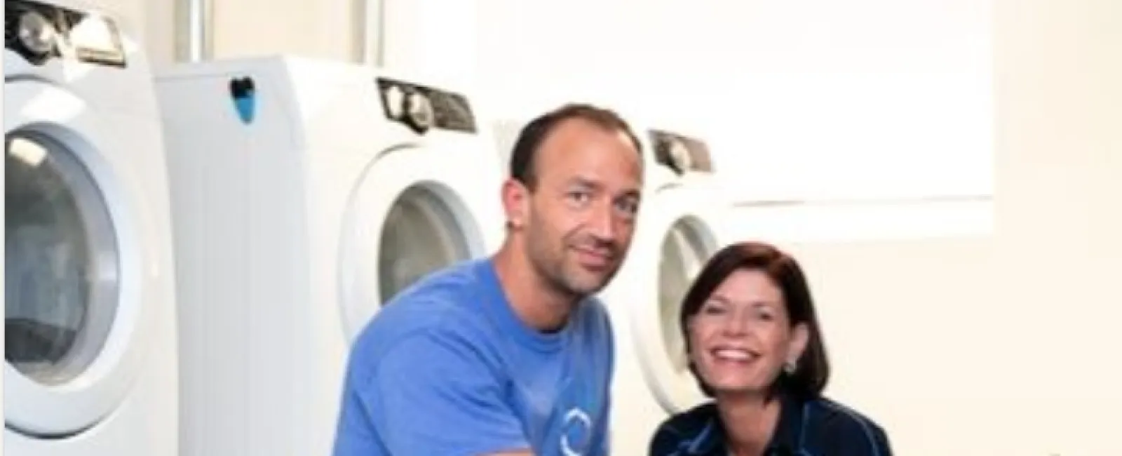 Two people in a laundry room folding clothes near stacked washers and dryers with cleaning supplies nearby.