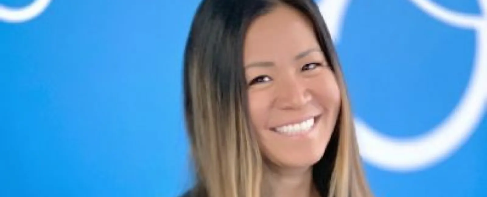 Smiling woman with long ombre hair wearing a black top standing against a blue wall with white circular patterns.
