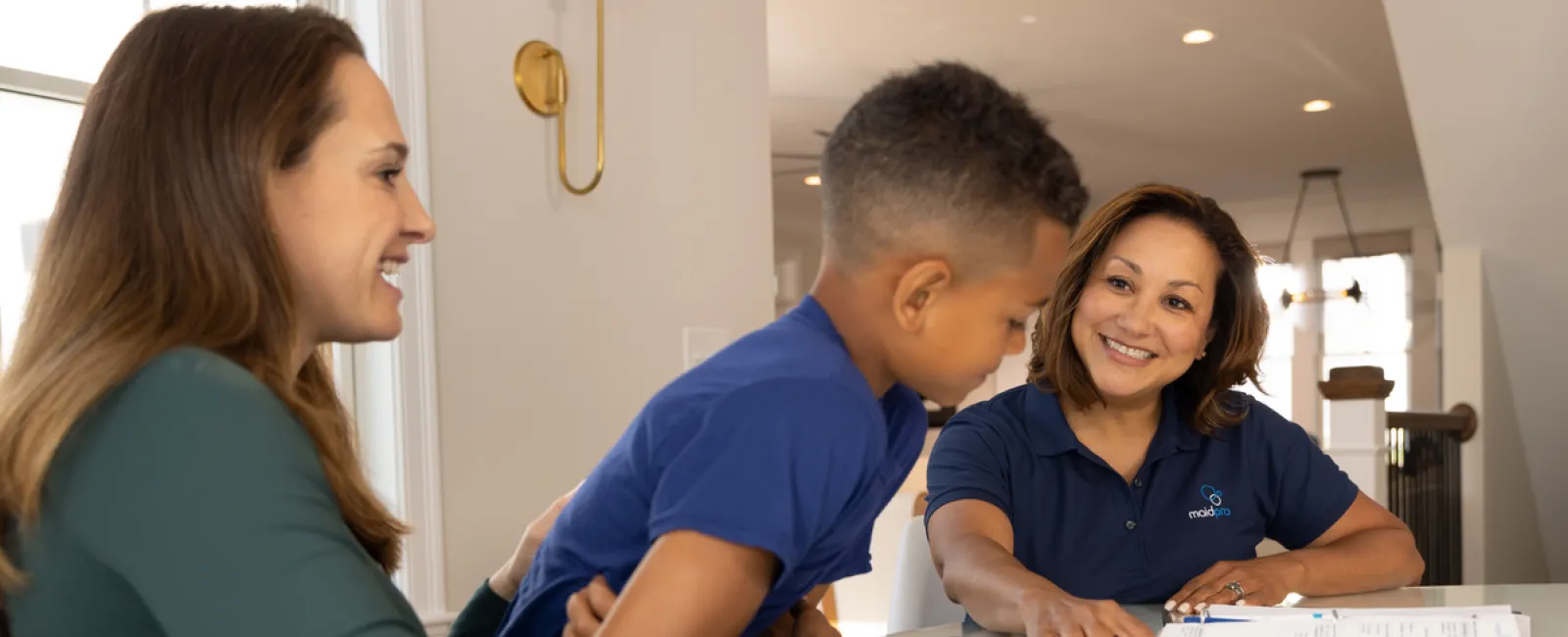 Tutor explaining homework to smiling boy and woman at a bright table in a modern home setting.