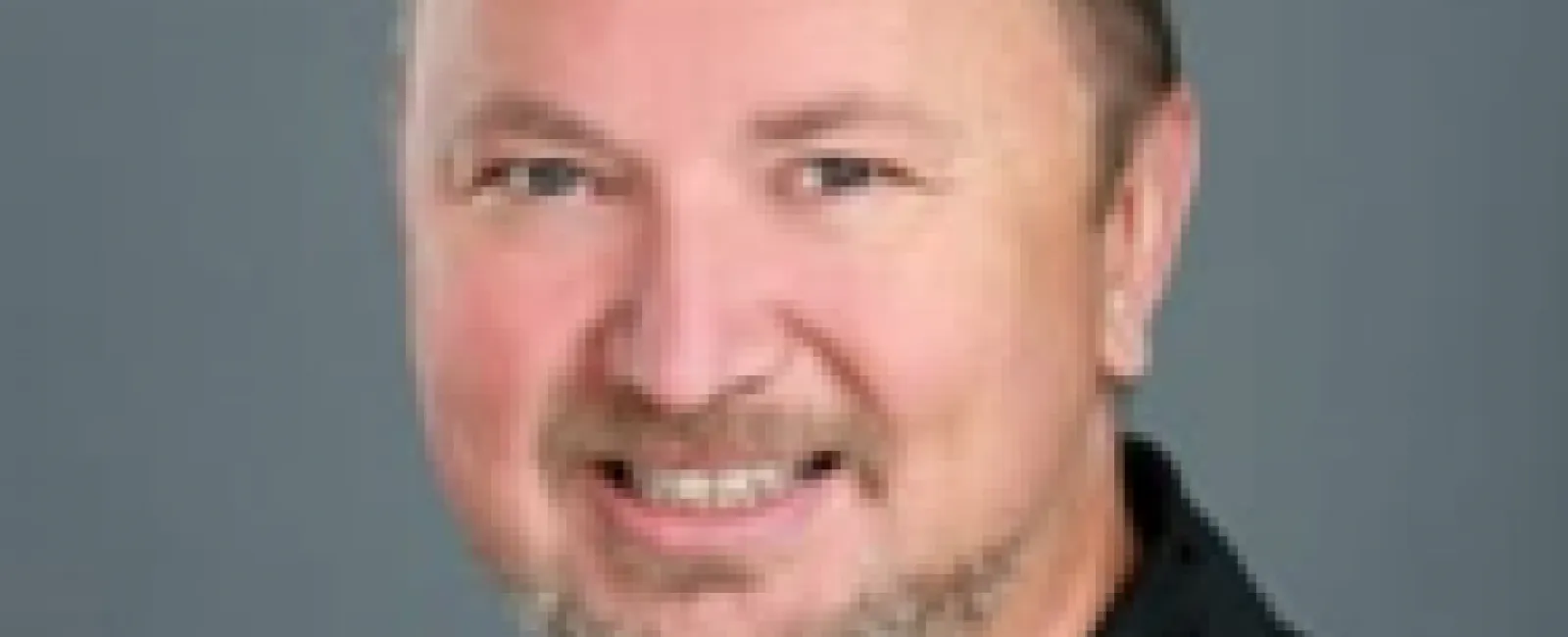 Portrait of a smiling man with short curly hair and a trimmed beard wearing a black zip-up jacket.